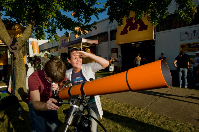 young man and woman looking through large orange telescope in front of U of M building at Minnesota State Fair