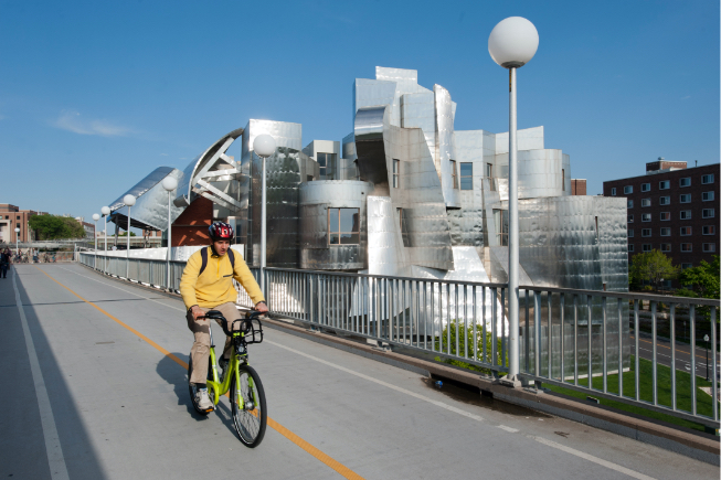 Student riding a NiceRide bicycle across pedestrian bridge with Weisman museum in background