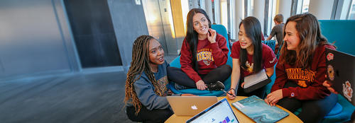 A student study group sits before laptops on a table in a modernly designed building
