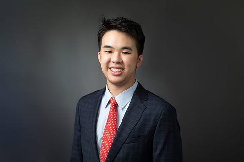Justin Ho smiles in front of a gray sunburst gradient background wearing a suit and tie