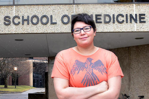 Goodsky, dark hair, dark glasses, with peach-colored T-shirt with phoenix graphic, stands, arms crossed, before a building labeled School of Medicine.